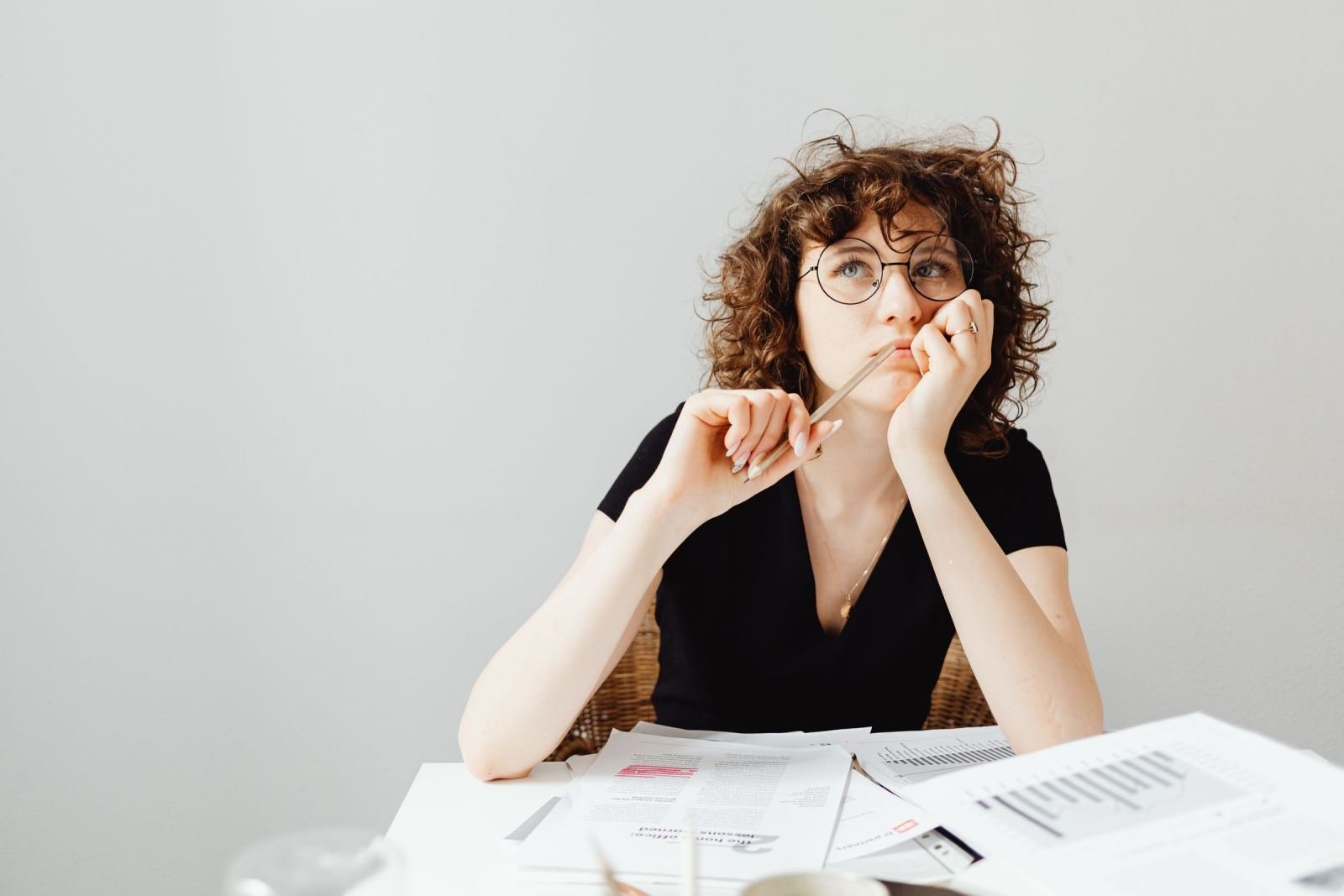 A woman with glasses sits at a table with papers and looks thoughtful.