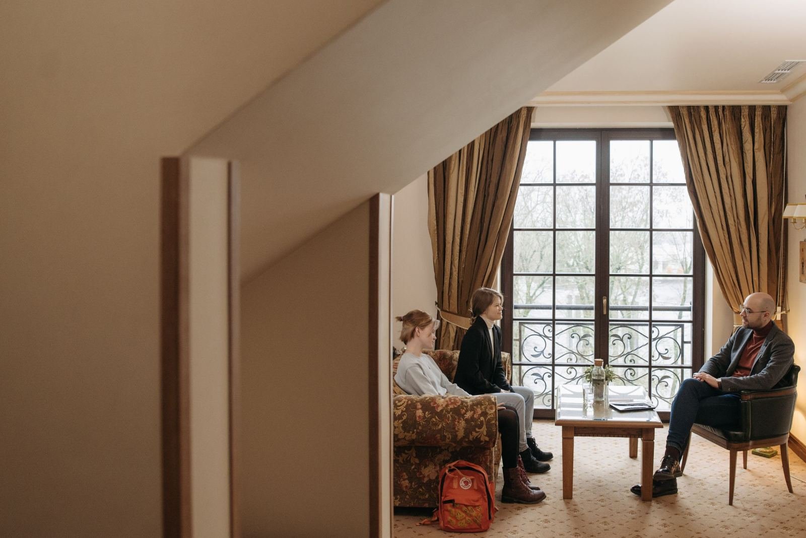 Three people sit in armchairs and talk in a bright room with a large window