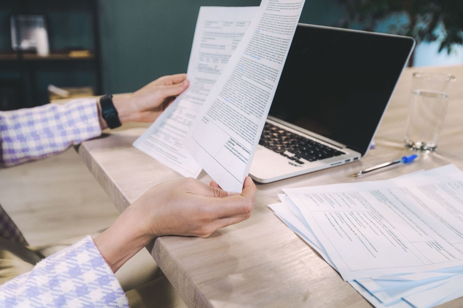 Two people review printed documents at a table with a laptop and a glass of water