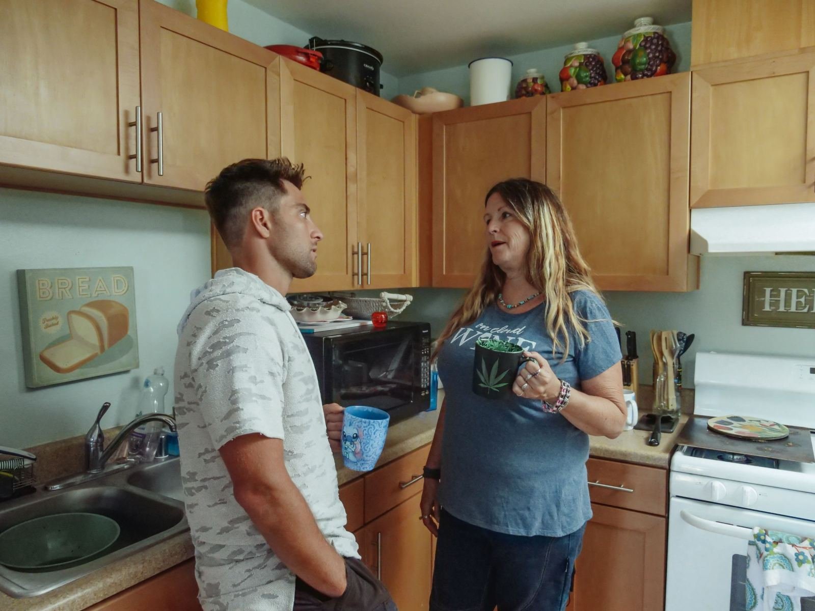 A man and a woman hold mugs and talk in a kitchen