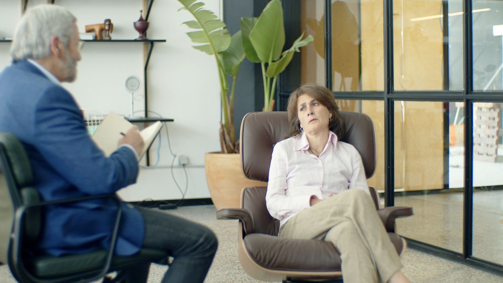 A man with a notebook talks to a woman sitting in a leather armchair