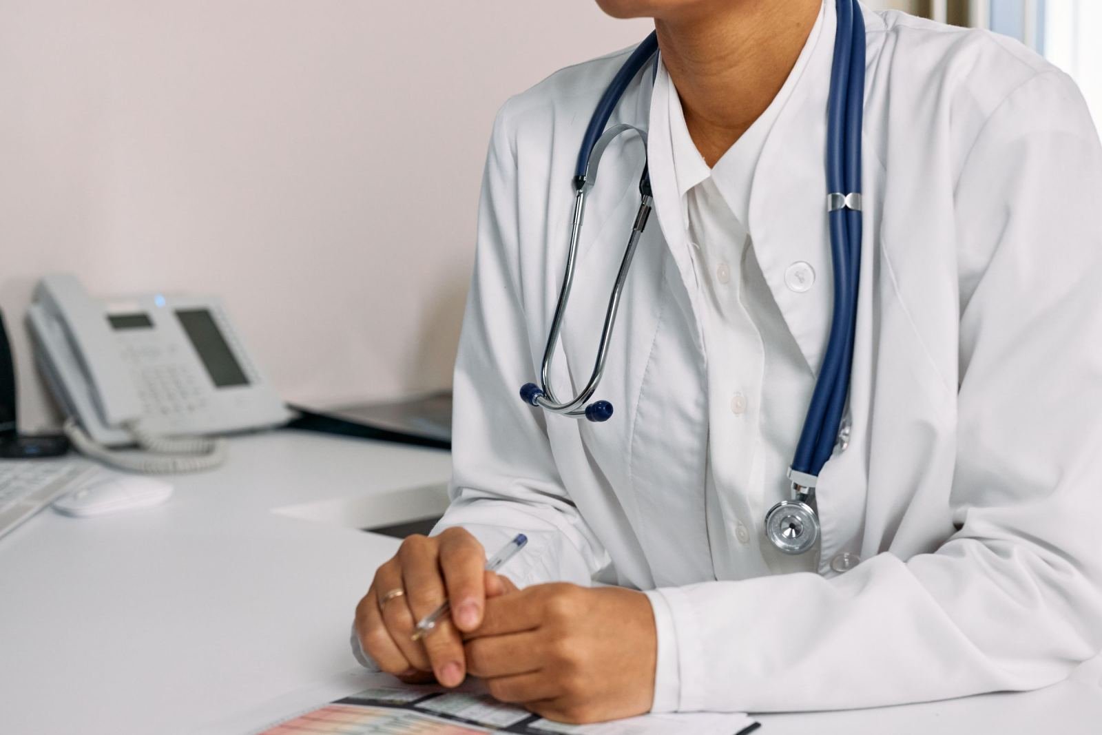 A clinician in a white lab coat and stethoscope sits at a desk with a pen