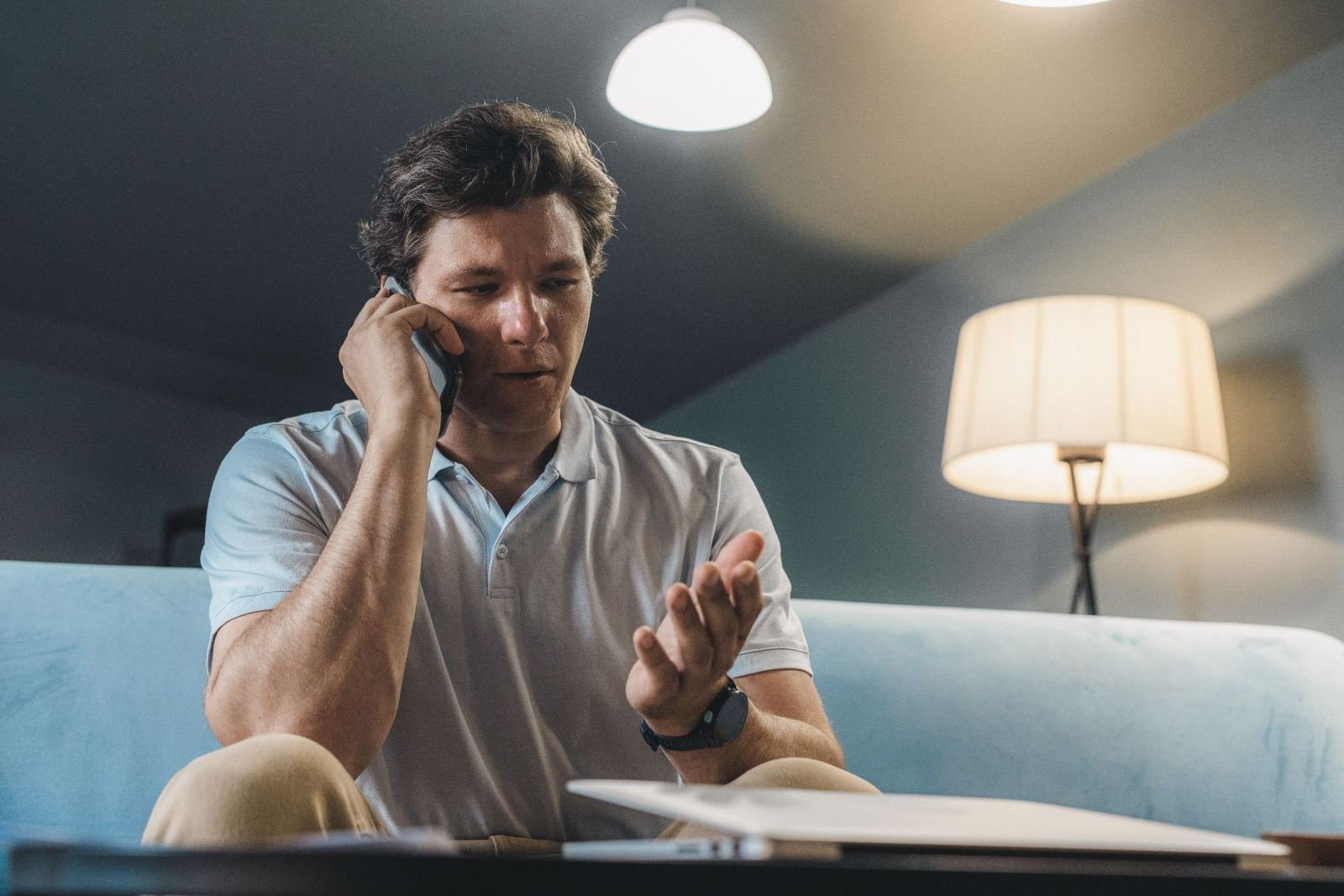 A seated man uses a cell phone. He is on a couch with a notebook close by.
