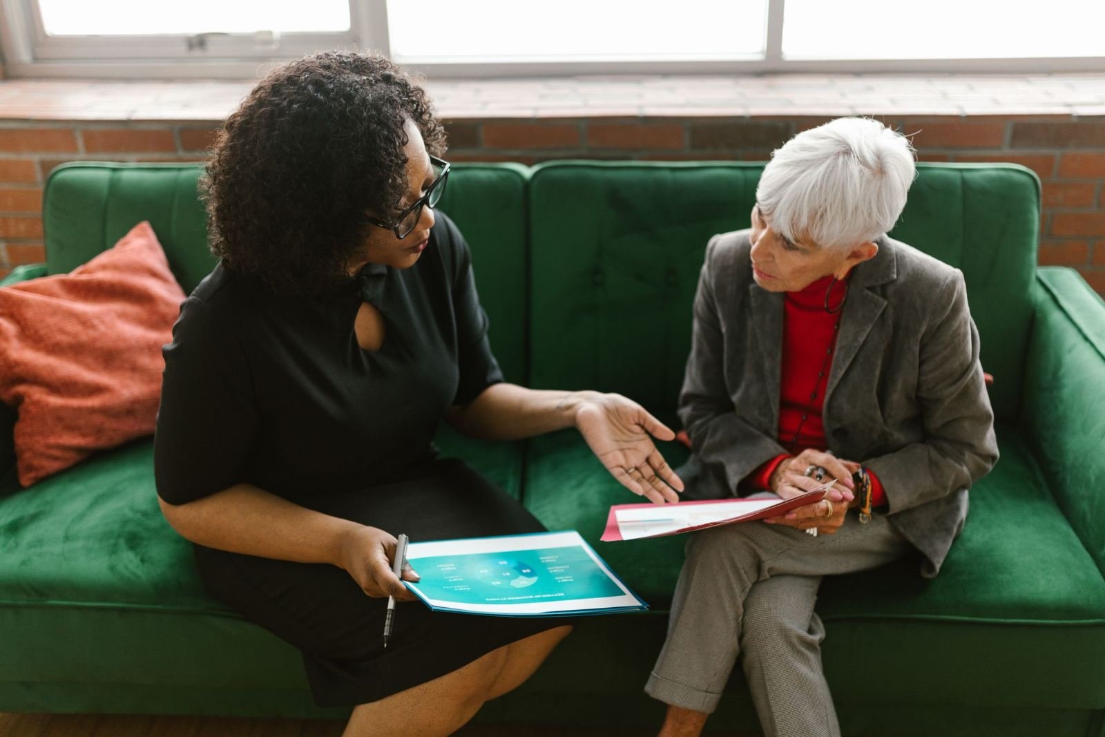 Two women sit on a green sofa reviewing documents together