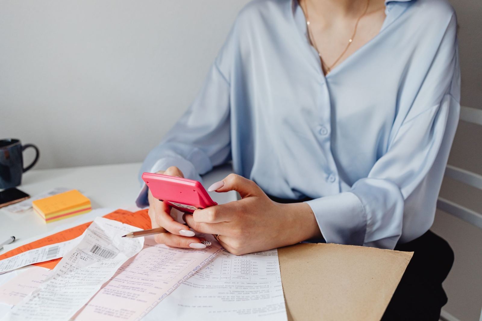 A person using a pink calculator to review printed receipts on a white desk