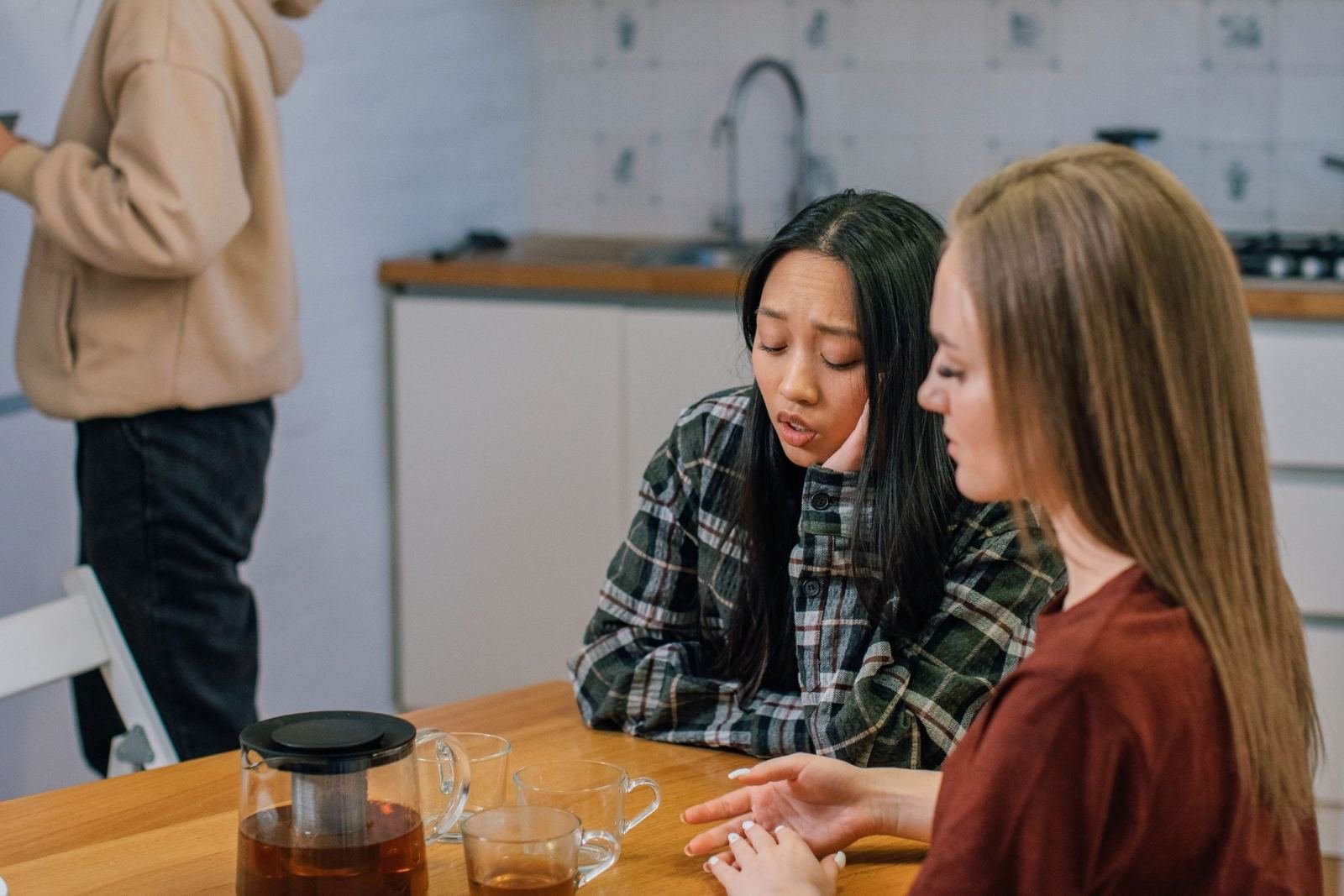 Two women talk at a kitchen table, one with a concerned expression