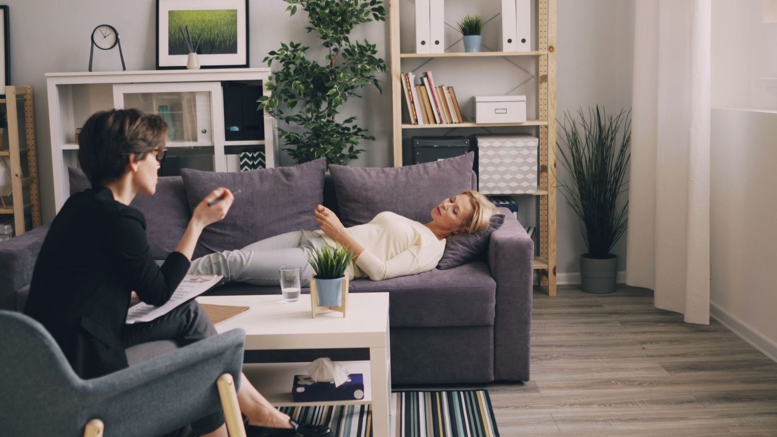 A woman lies back on a gray couch while a counselor in a dark jacket speaks with her