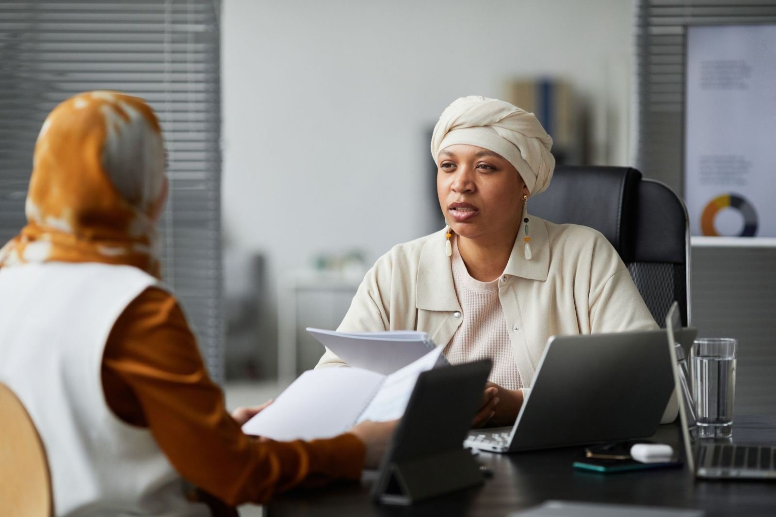 Two women sit at a desk in a bright office with papers and a laptop.
