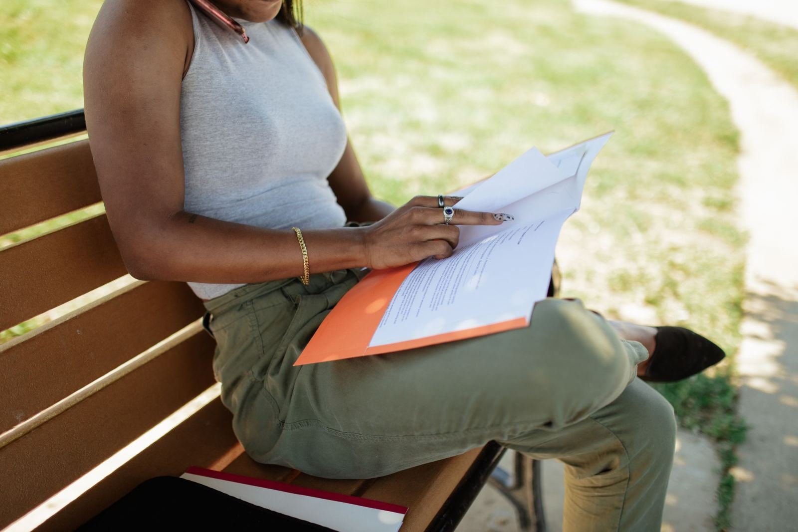 Someone sits on a park bench reading a paper with an orange cover.