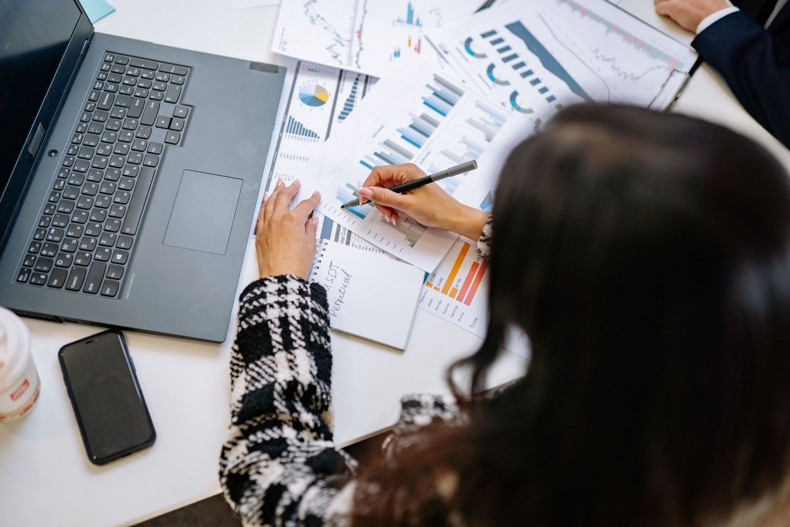 A person uses a pen to review financial charts and graphs on a white desk next to a laptop