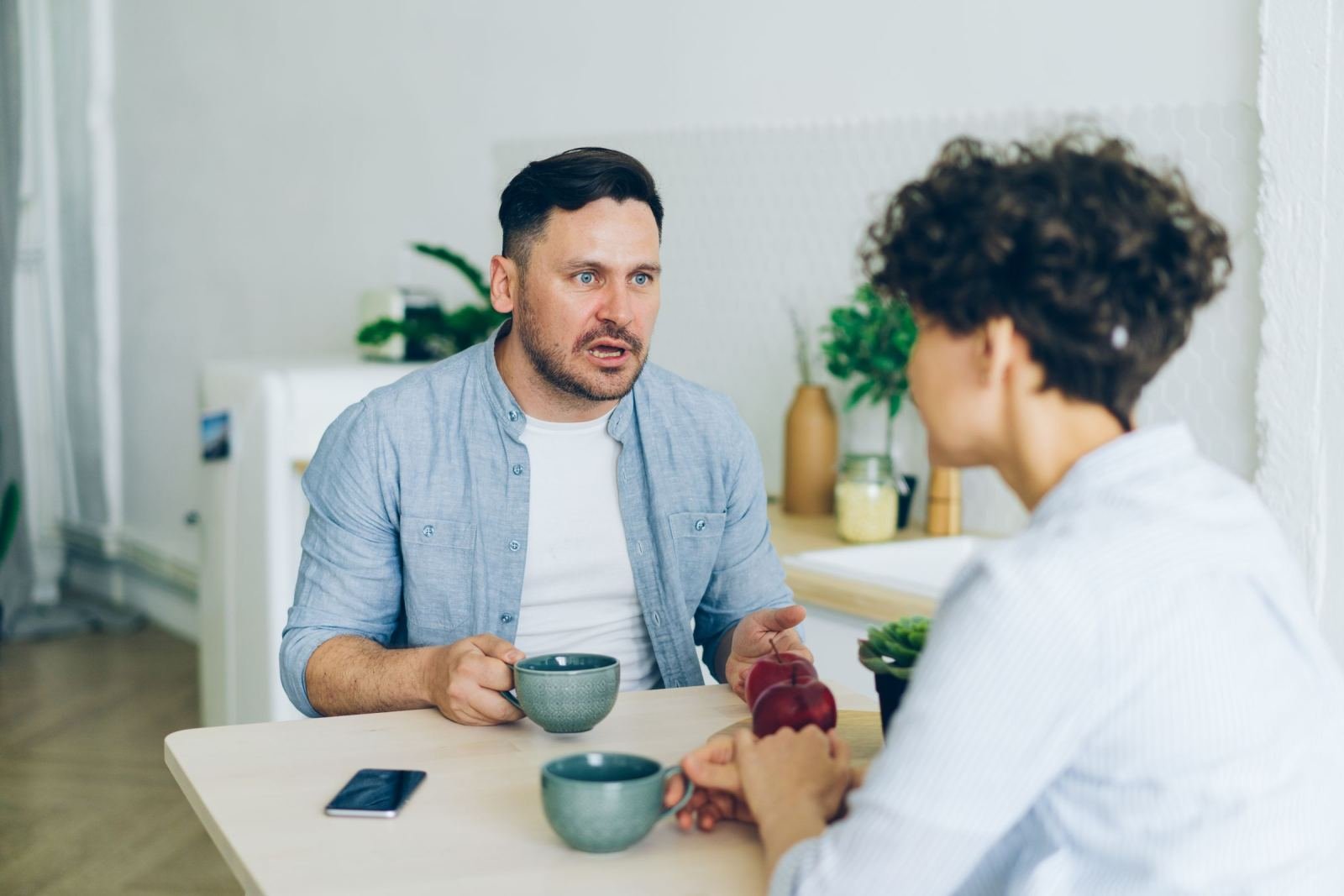 A man and woman talk across a table with coffee mugs and apples