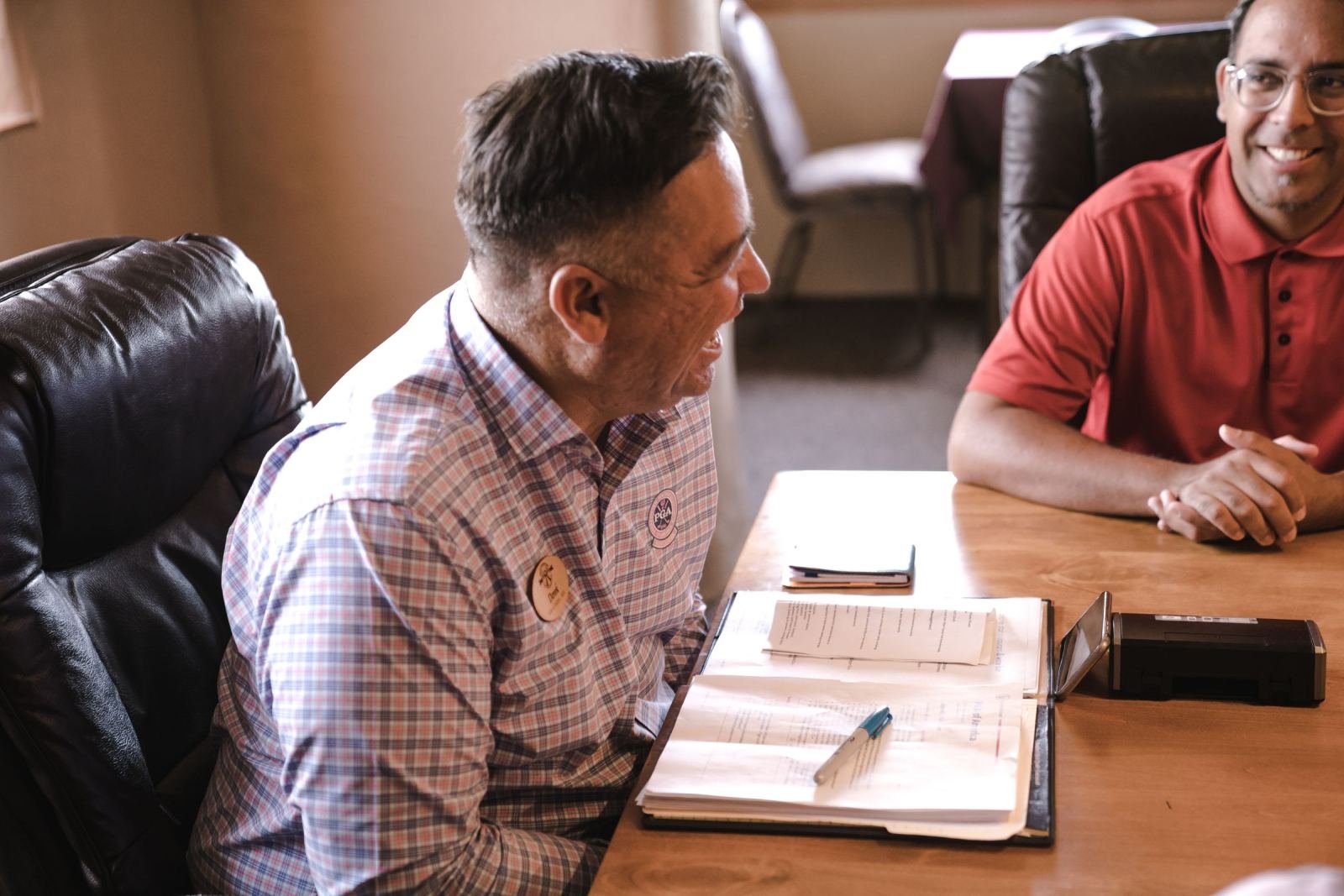 Two men talk about papers at a desk in a bright office.
