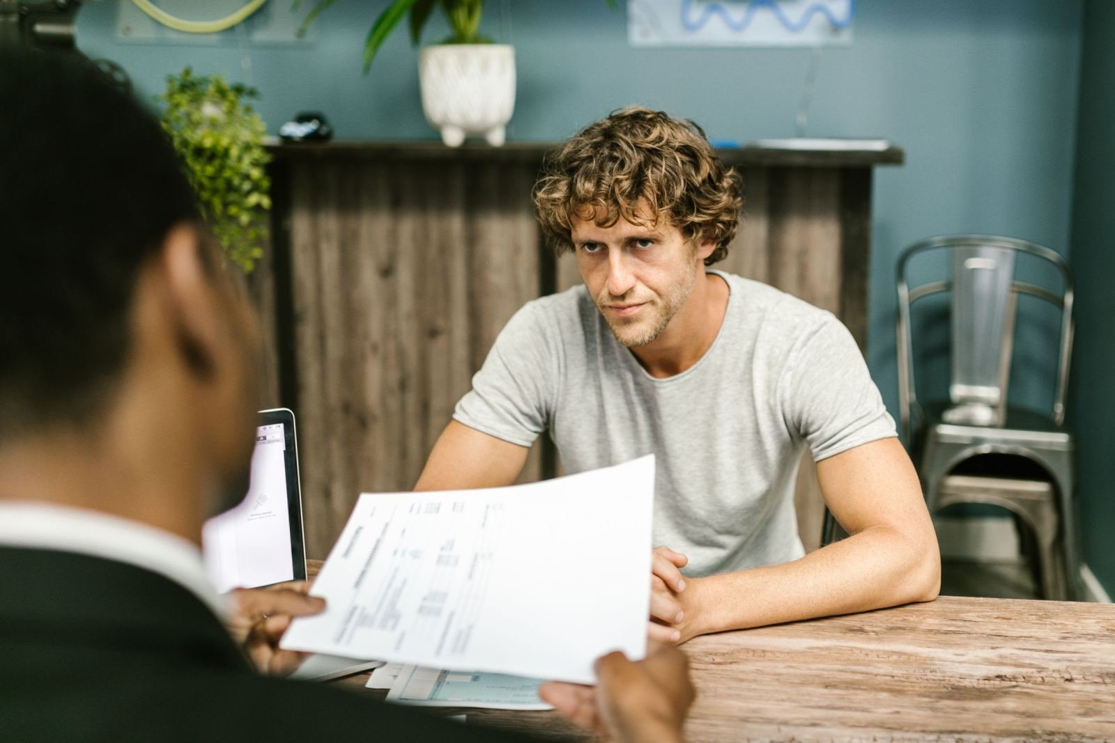 A professional holds a paper while a man in a grey shirt looks at it across a desk.