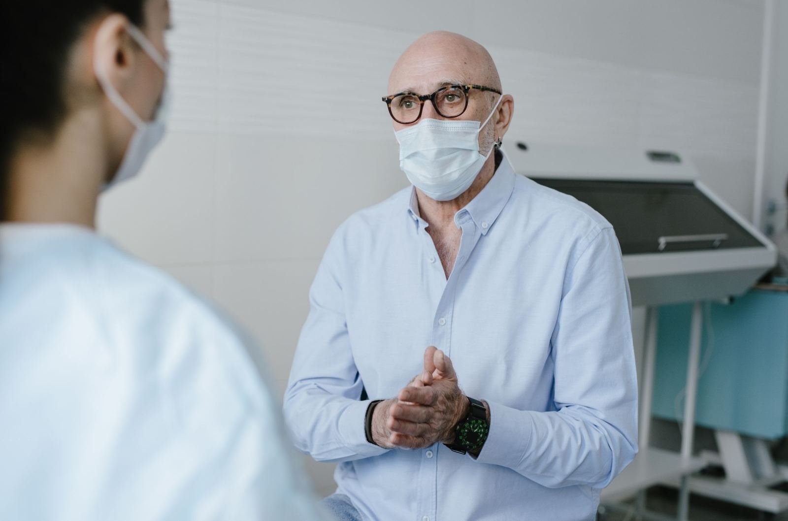 A man in a surgical mask and glasses speaks with a healthcare provider