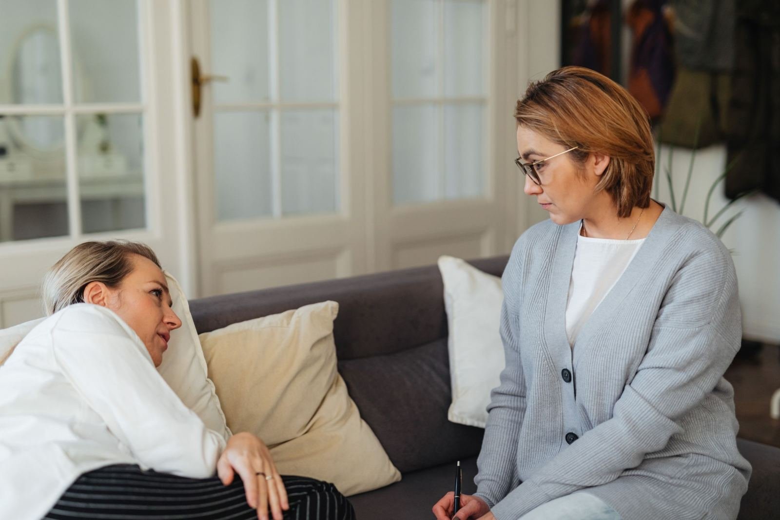 One woman leans on a couch while the other holds a pen. They chat in a well-lit room.
