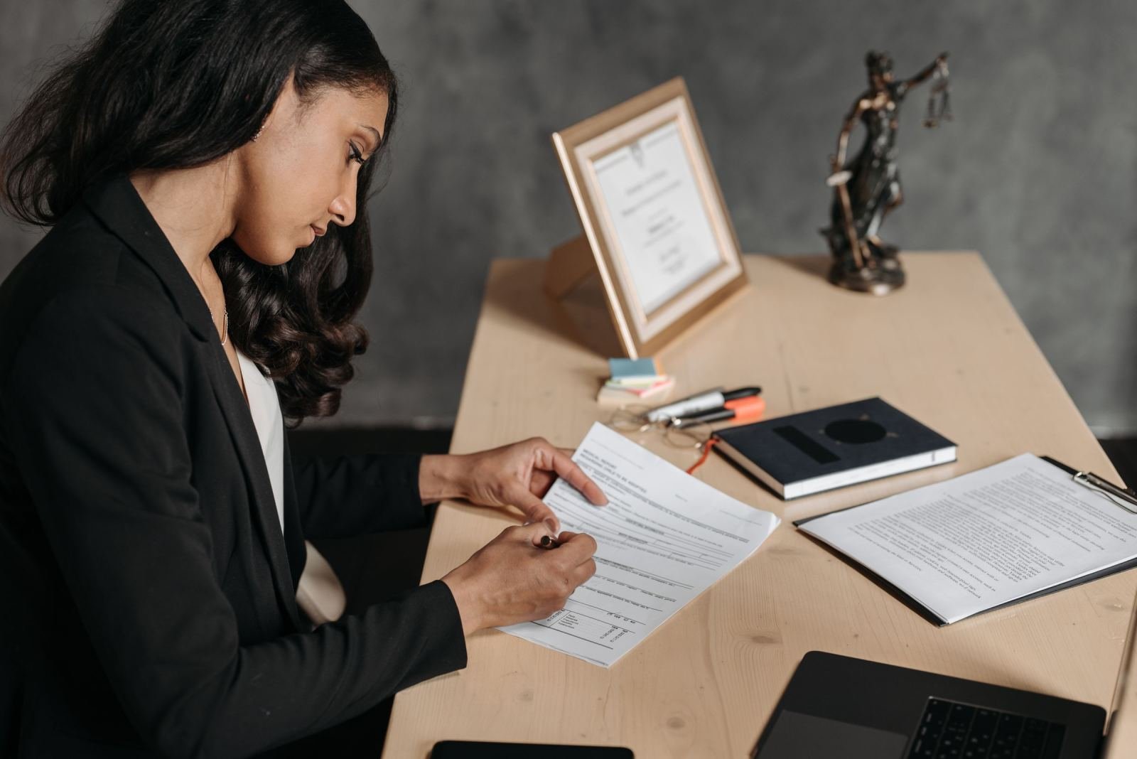 A woman in a blazer reviews a document at a desk with a statue of Lady Justice
