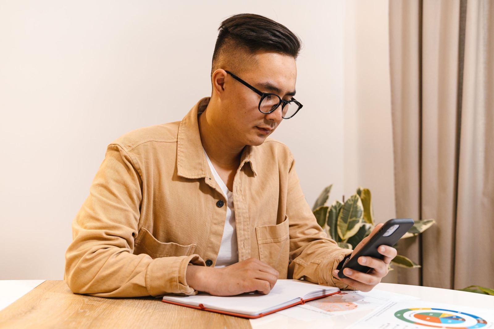 A man wearing glasses checks his phone at a table. He has a notebook in front of him.