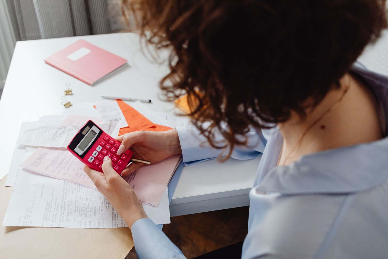 A person using a red calculator while reviewing papers at a white desk