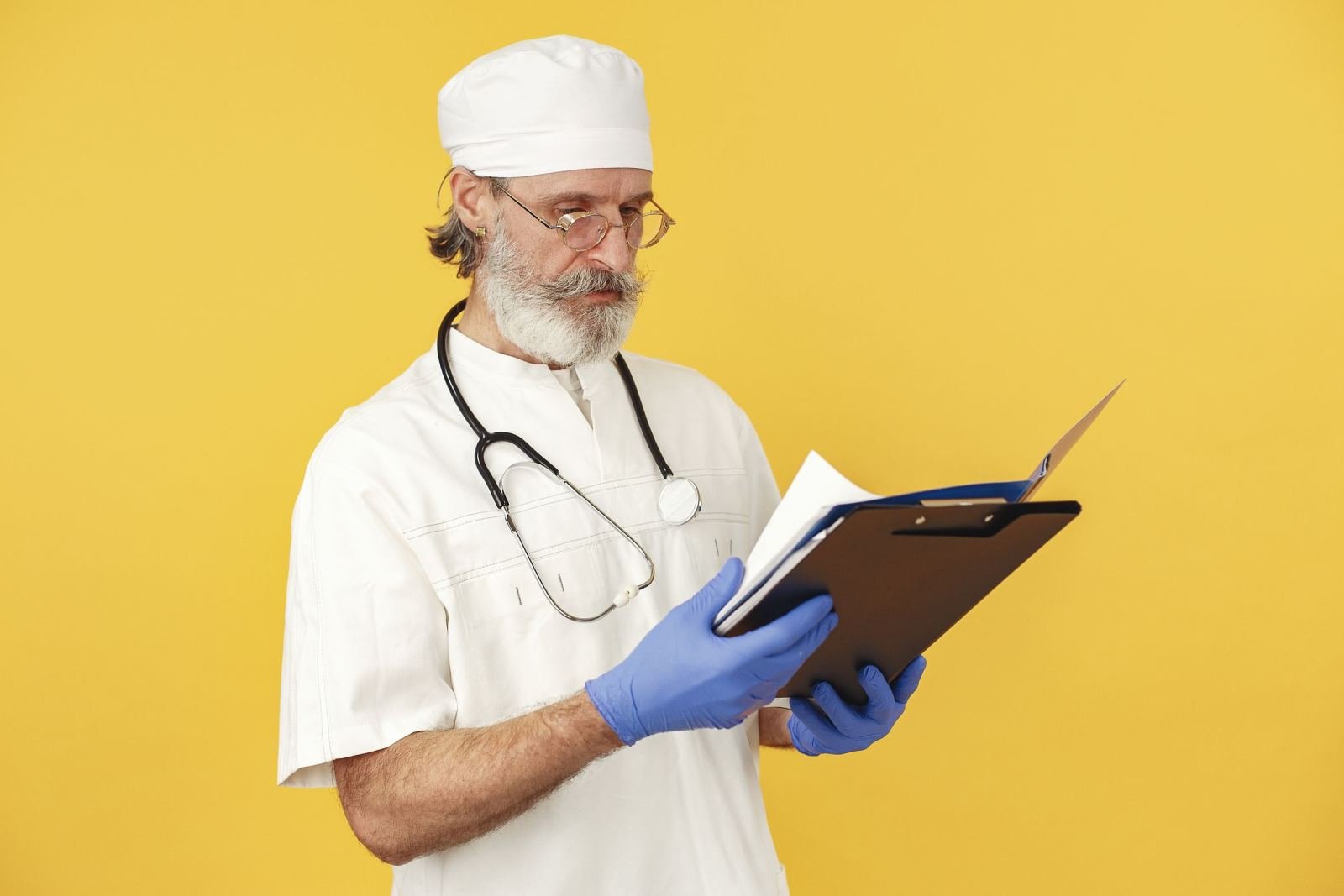 A doctor in a white coat and blue gloves looks at a clipboard.