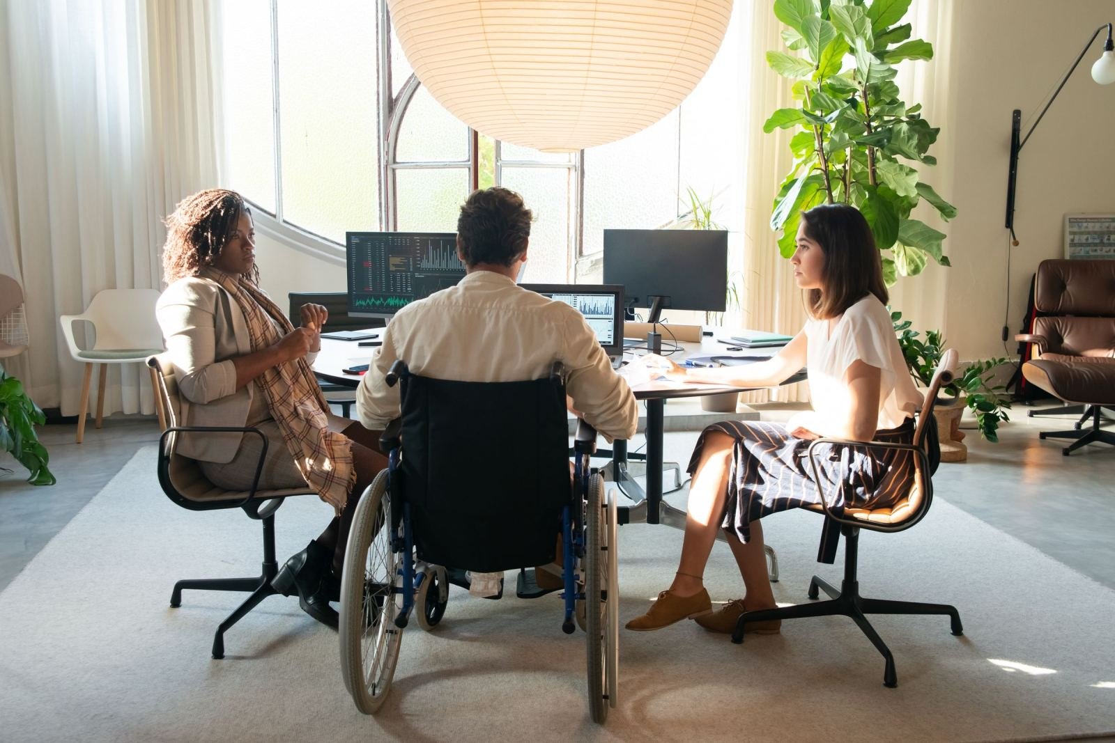 Two women and a person using a wheelchair sit at a desk with computer screens.
