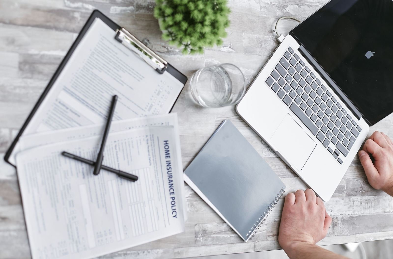 A laptop, clipboard, and notebook on a desk with a person's hands visible