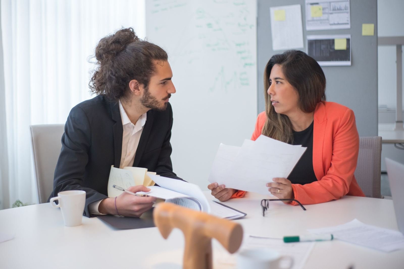 A man and woman in professional attire discuss documents at a white table