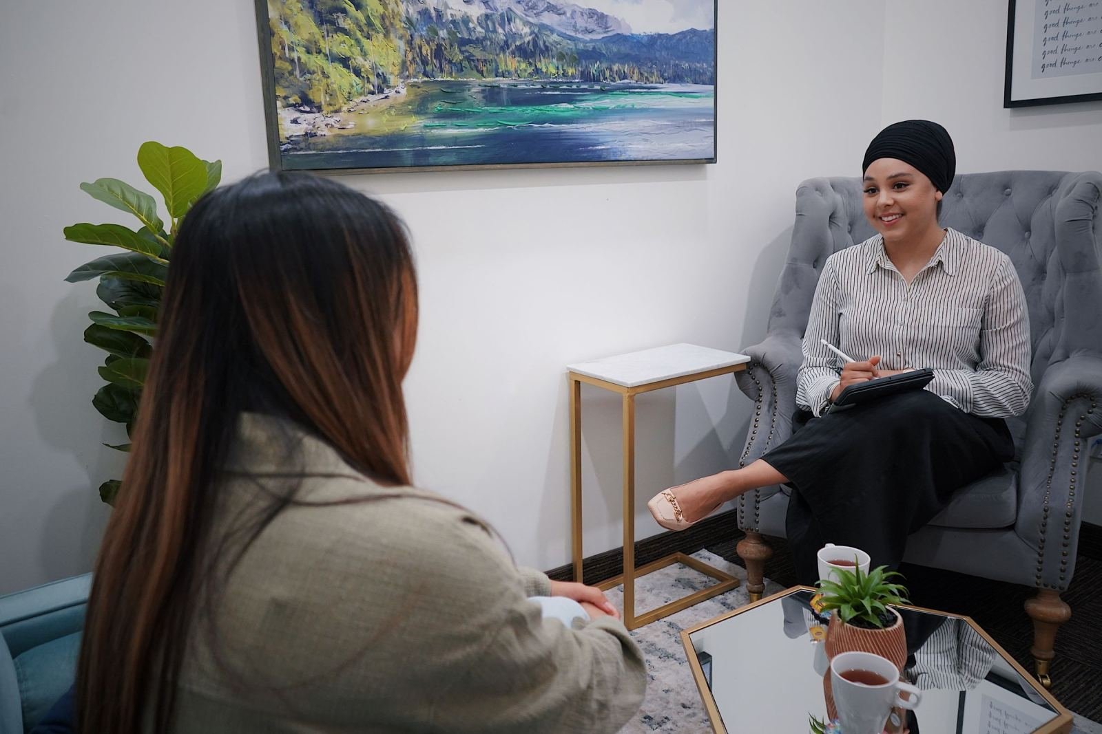 A smiling woman wearing a headscarf sits in a gray armchair. She is speaking with someone else.
