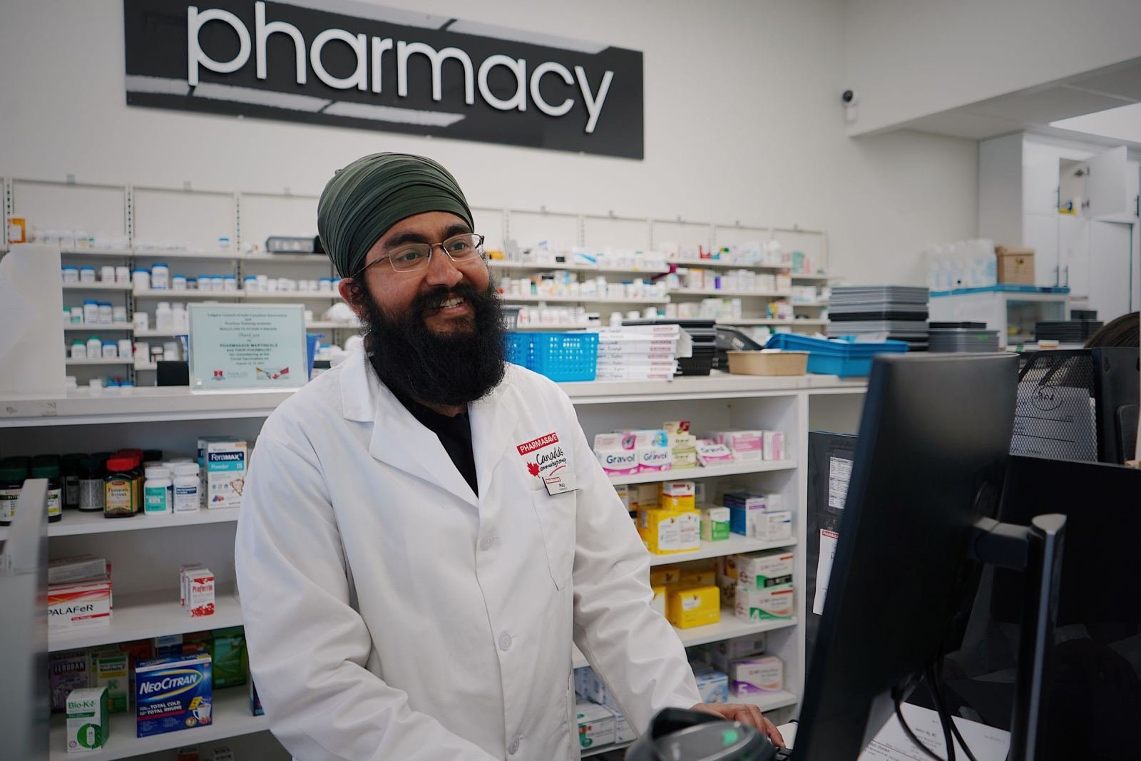 A happy pharmacist wearing a white coat and a green turban stands behind the counter.