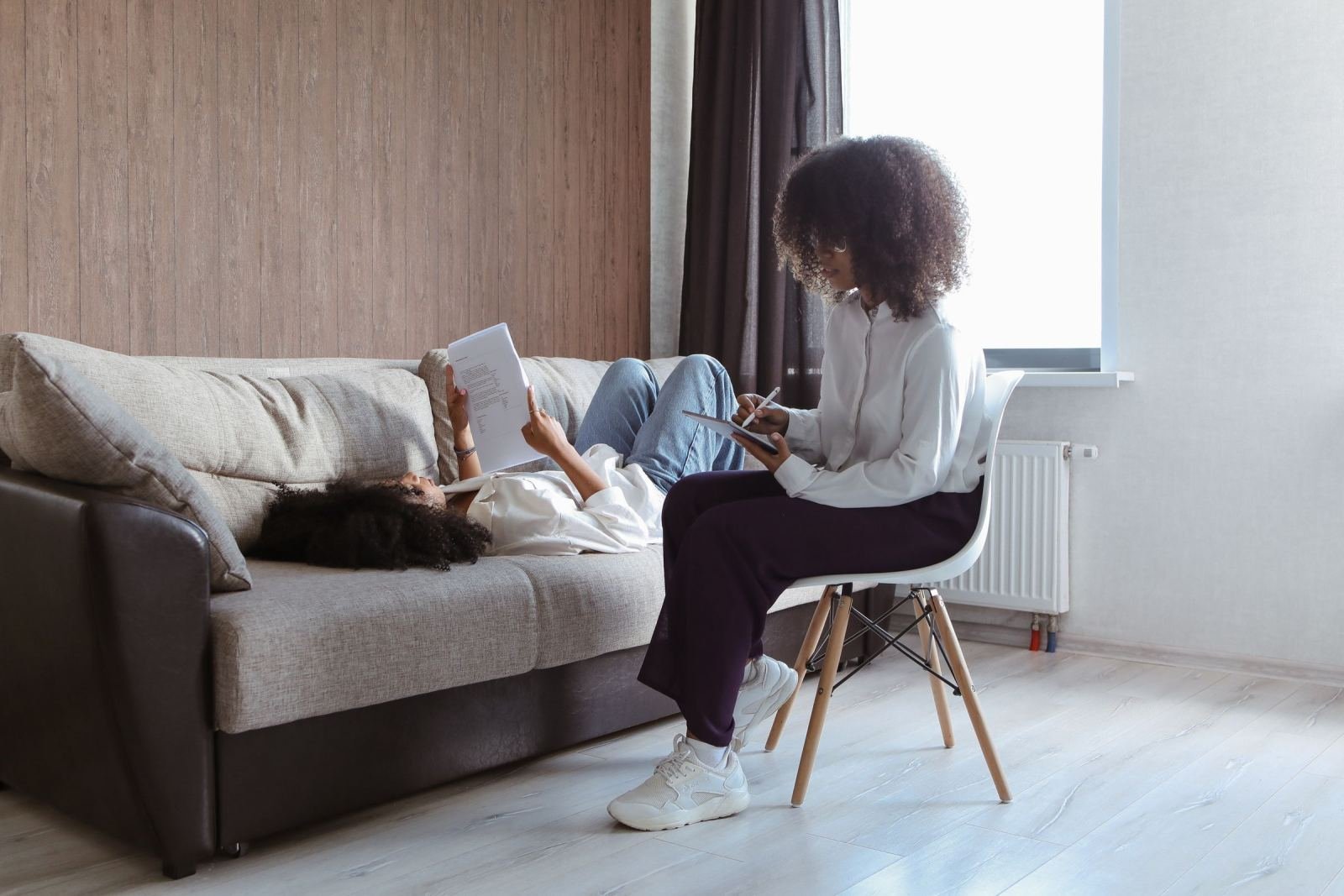 A woman sits in a chair taking notes while another woman lies on a sofa holding a paper