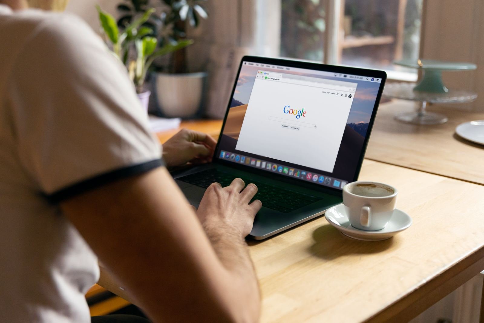 Someone sits at a wooden table with coffee, using a laptop to look up information online.