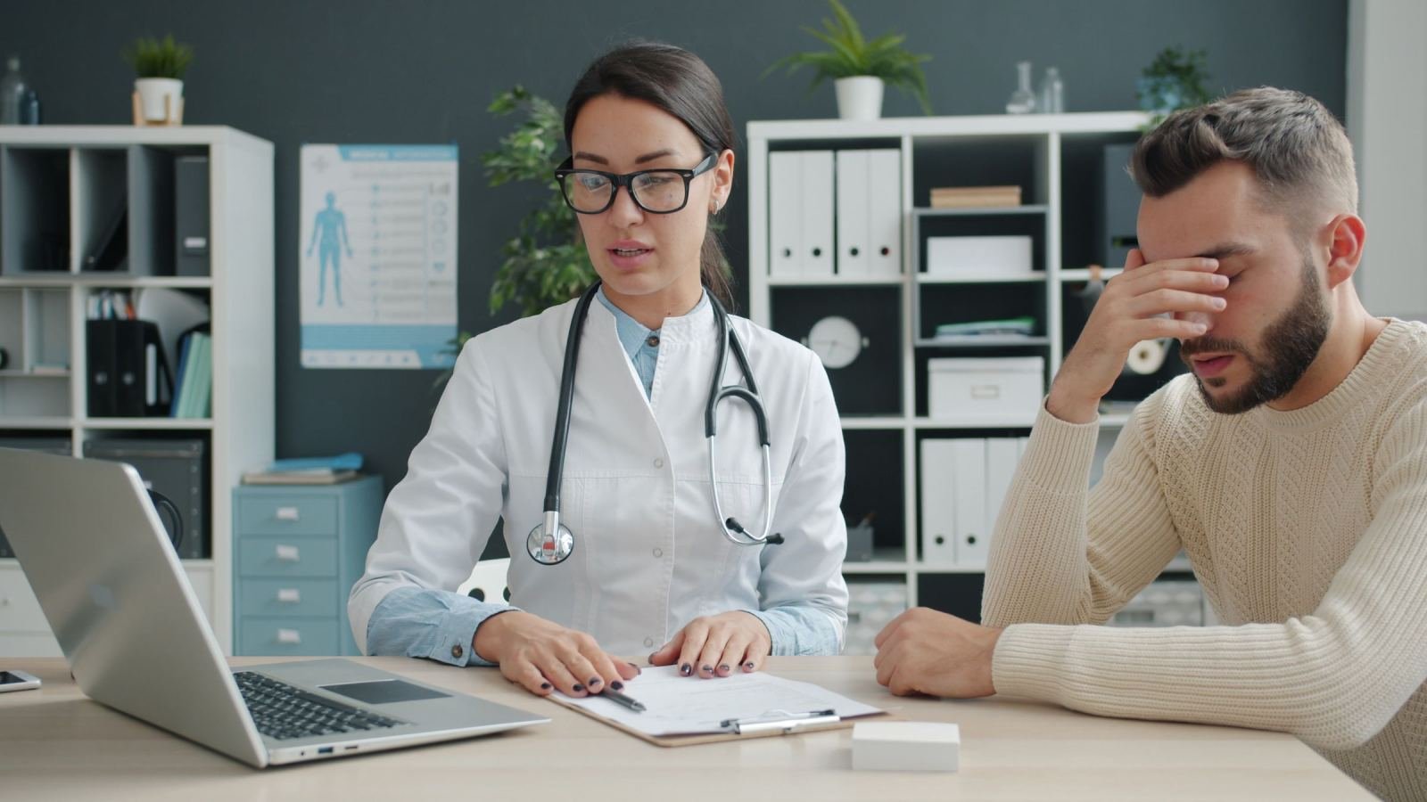 A healthcare provider in a white coat talks with a man at a desk