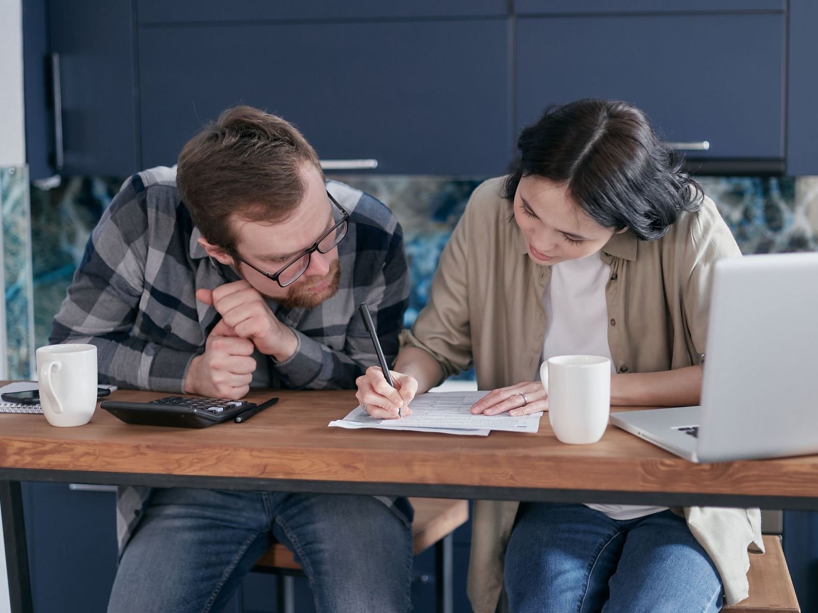 A couple looks over a paper form while sitting at a kitchen table with a laptop open.