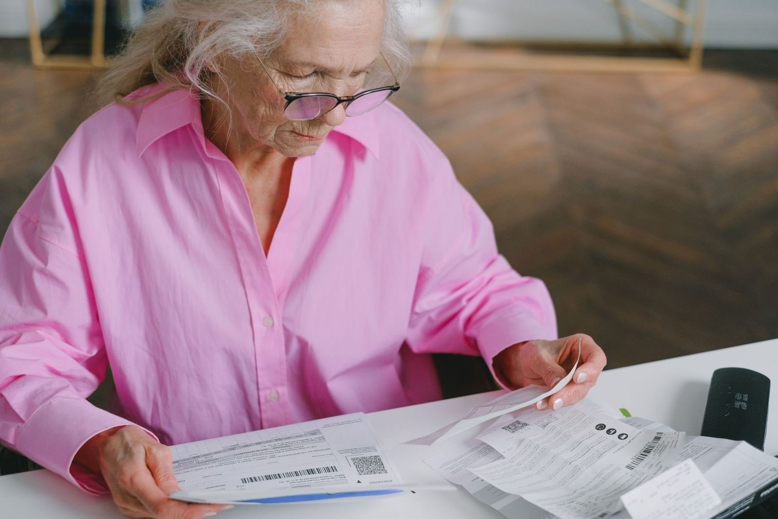 A senior lady in a pink top looks over forms on a white desk.