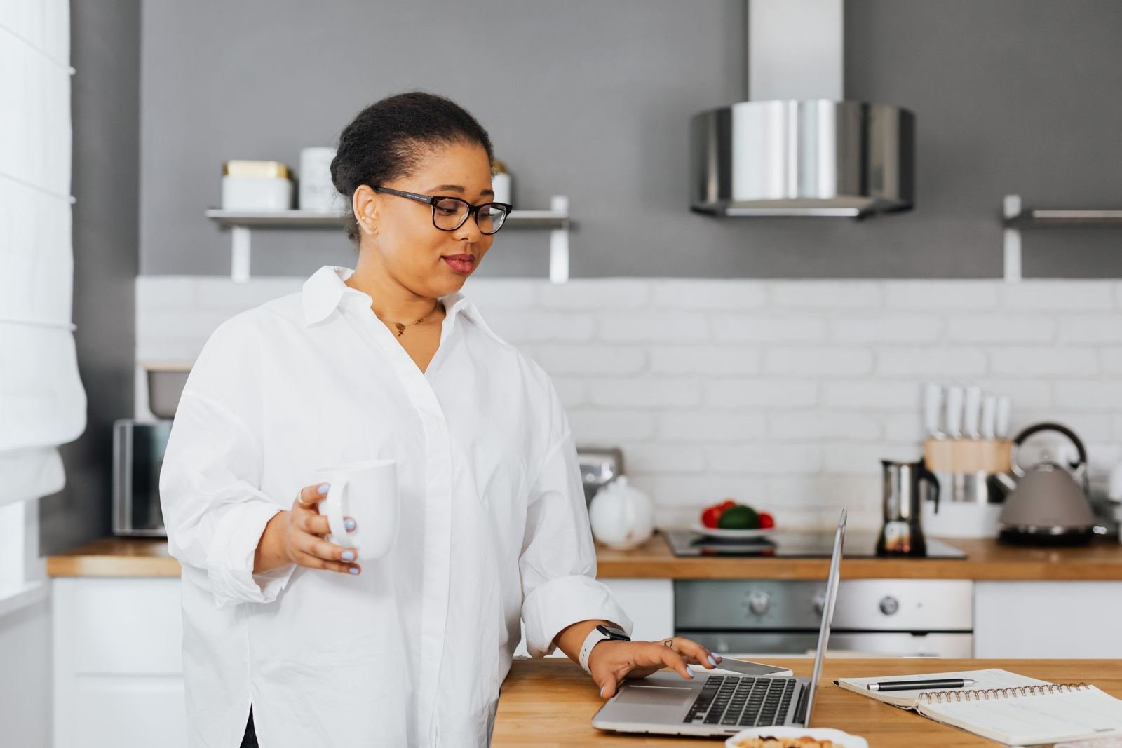 A woman in a white shirt sits in her kitchen. She uses a laptop and holds a mug.