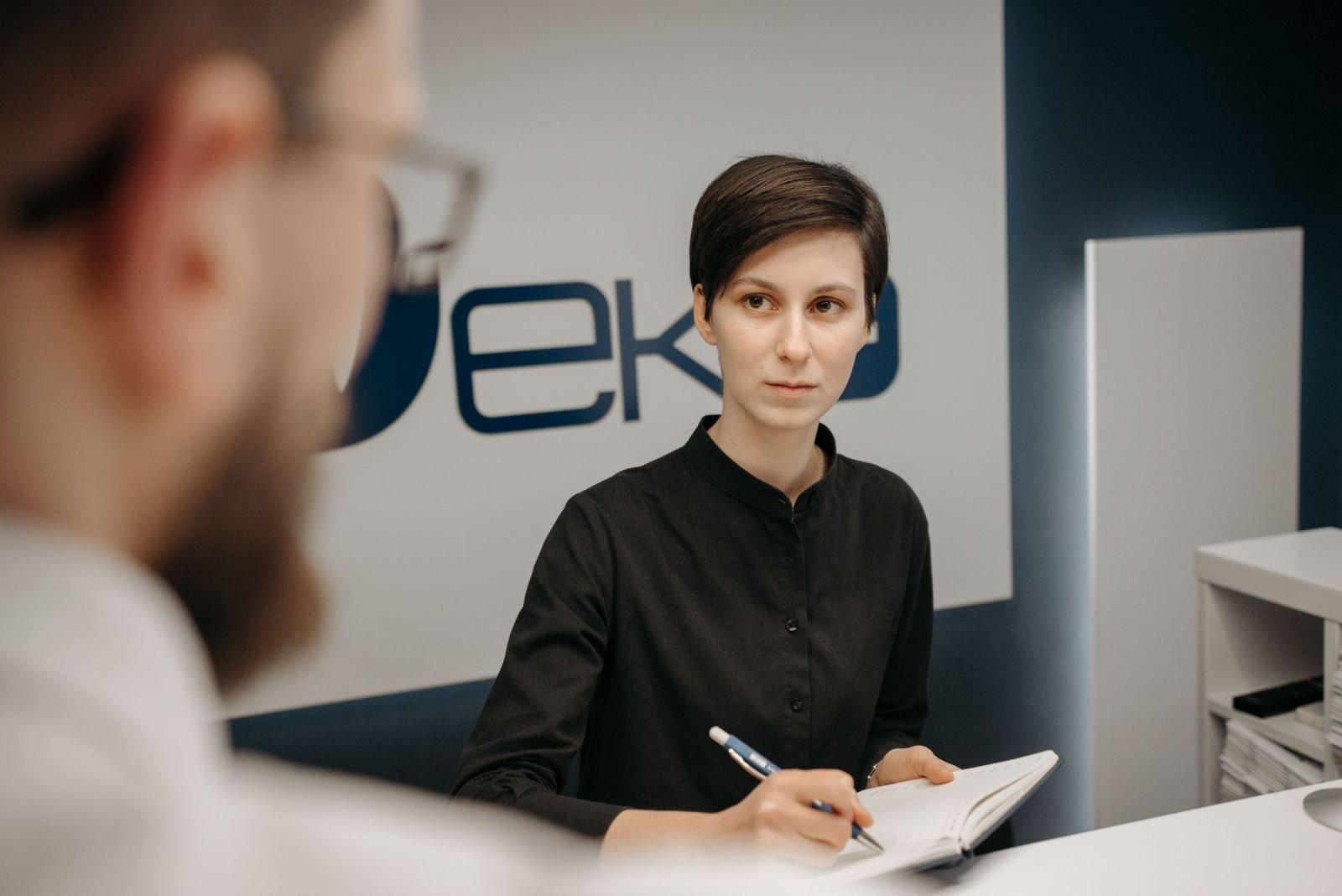 A woman at a reception desk holds a pen and notebook while speaking with a man