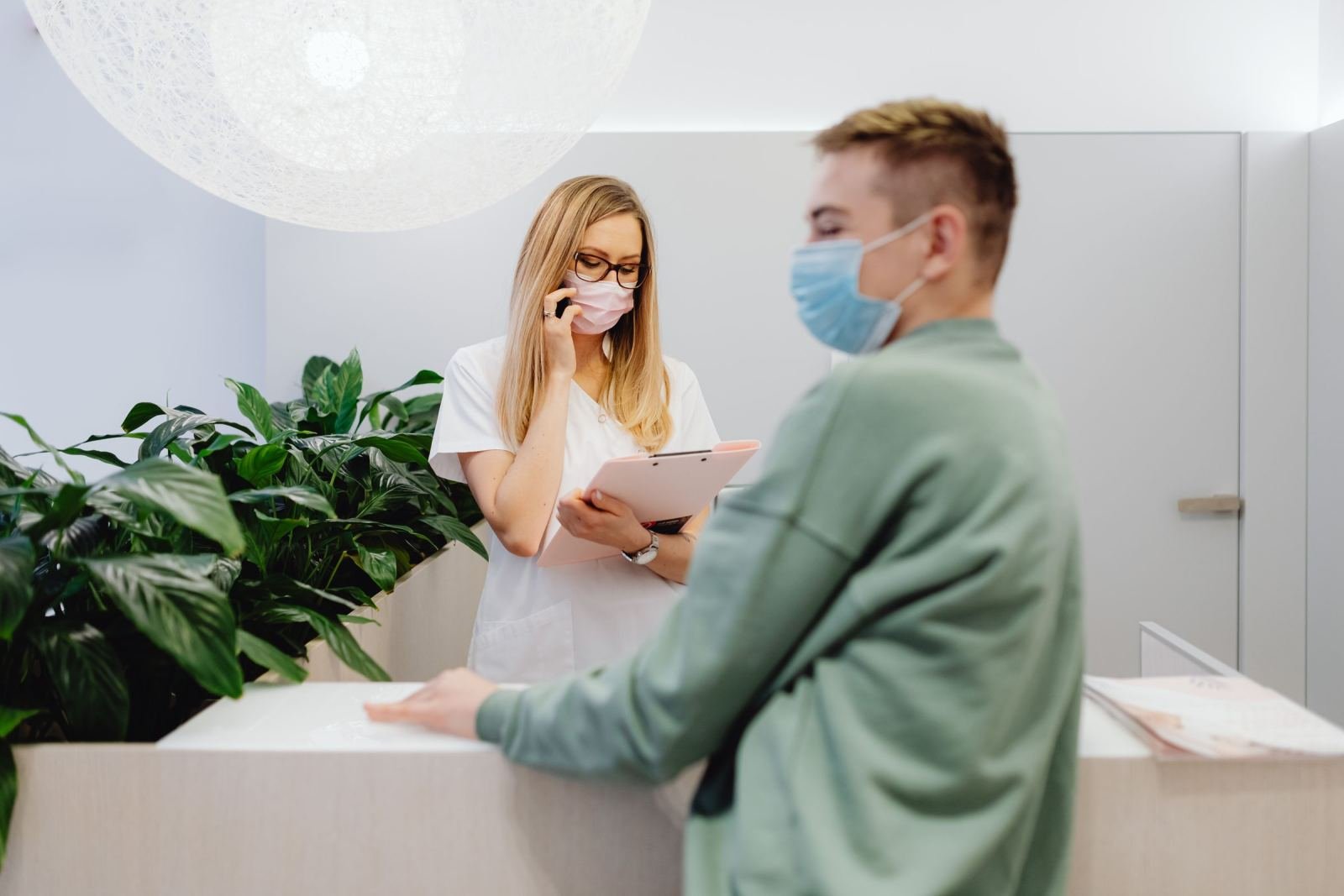 A medical receptionist in a mask speaks on the phone while attending to a patient