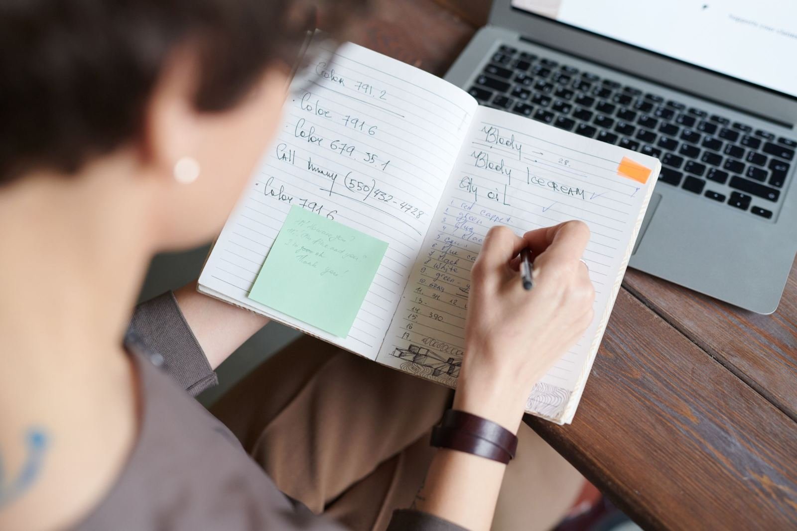 A person writes in a notebook beside a laptop on a wooden desk.