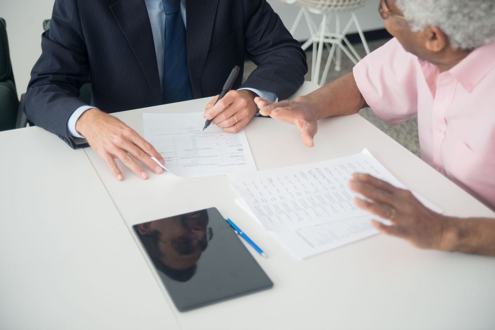 A man in a pink shirt talks with a professional in a suit. They sit across a desk covered in papers.
