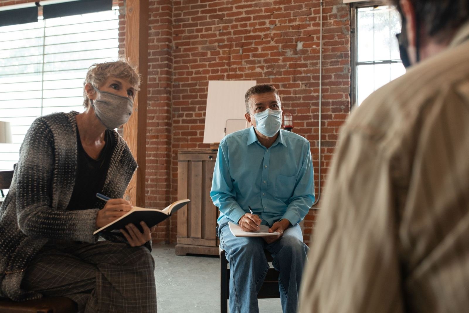 Three people meet in a brick room. Two wear face masks and hold notebooks while talking to the third person.