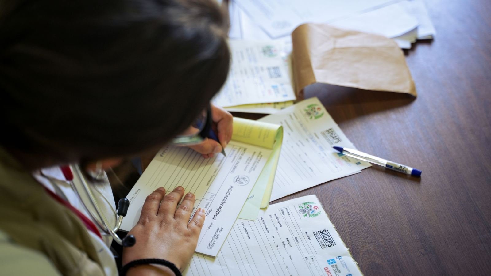 A healthcare provider in a white coat writes on a medical form at a desk