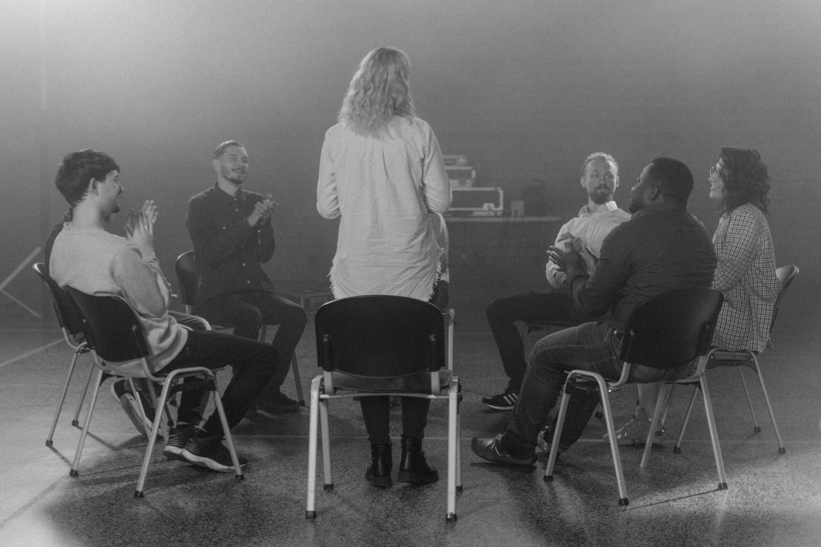 A group of adults sitting in a circle of chairs and listening to a person standing in the center