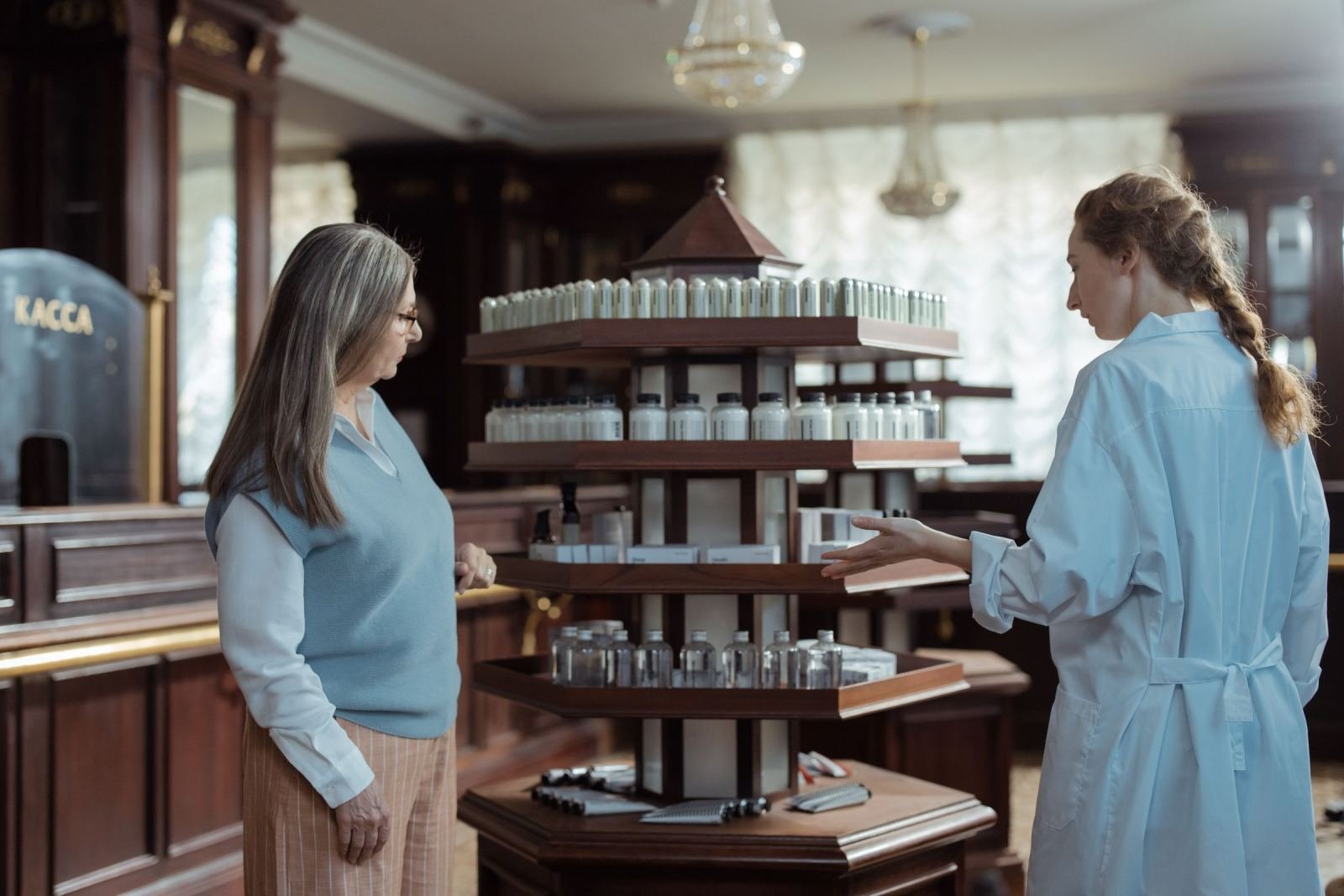 A pharmacist in a white coat talks with a customer next to a display of medicine bottles