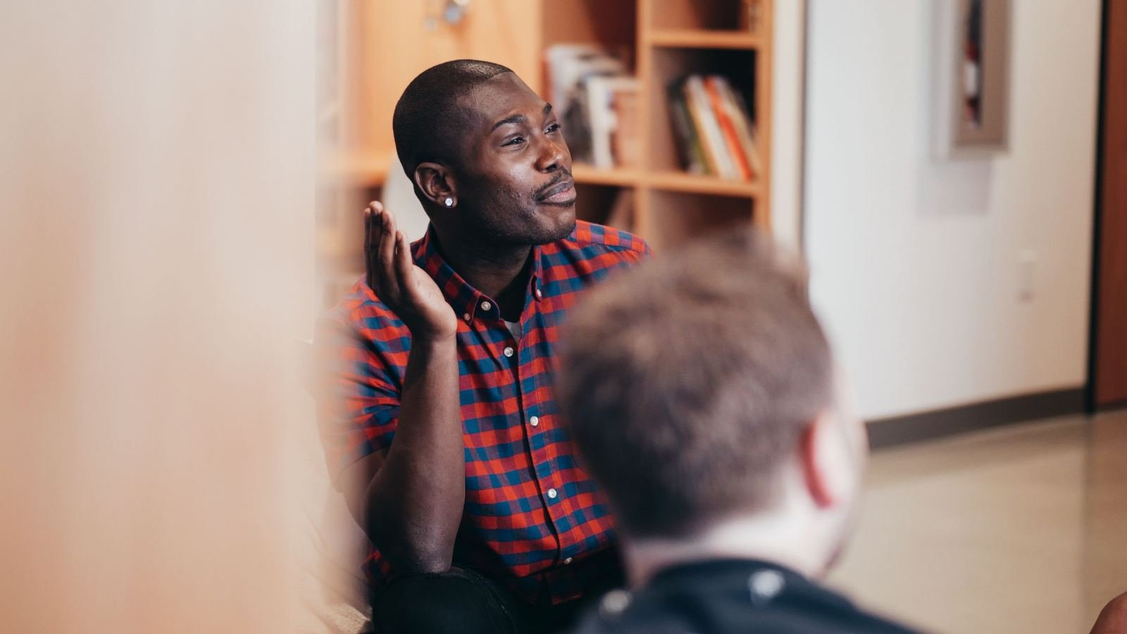 A man in a plaid shirt makes hand movements while speaking to another person in an office.