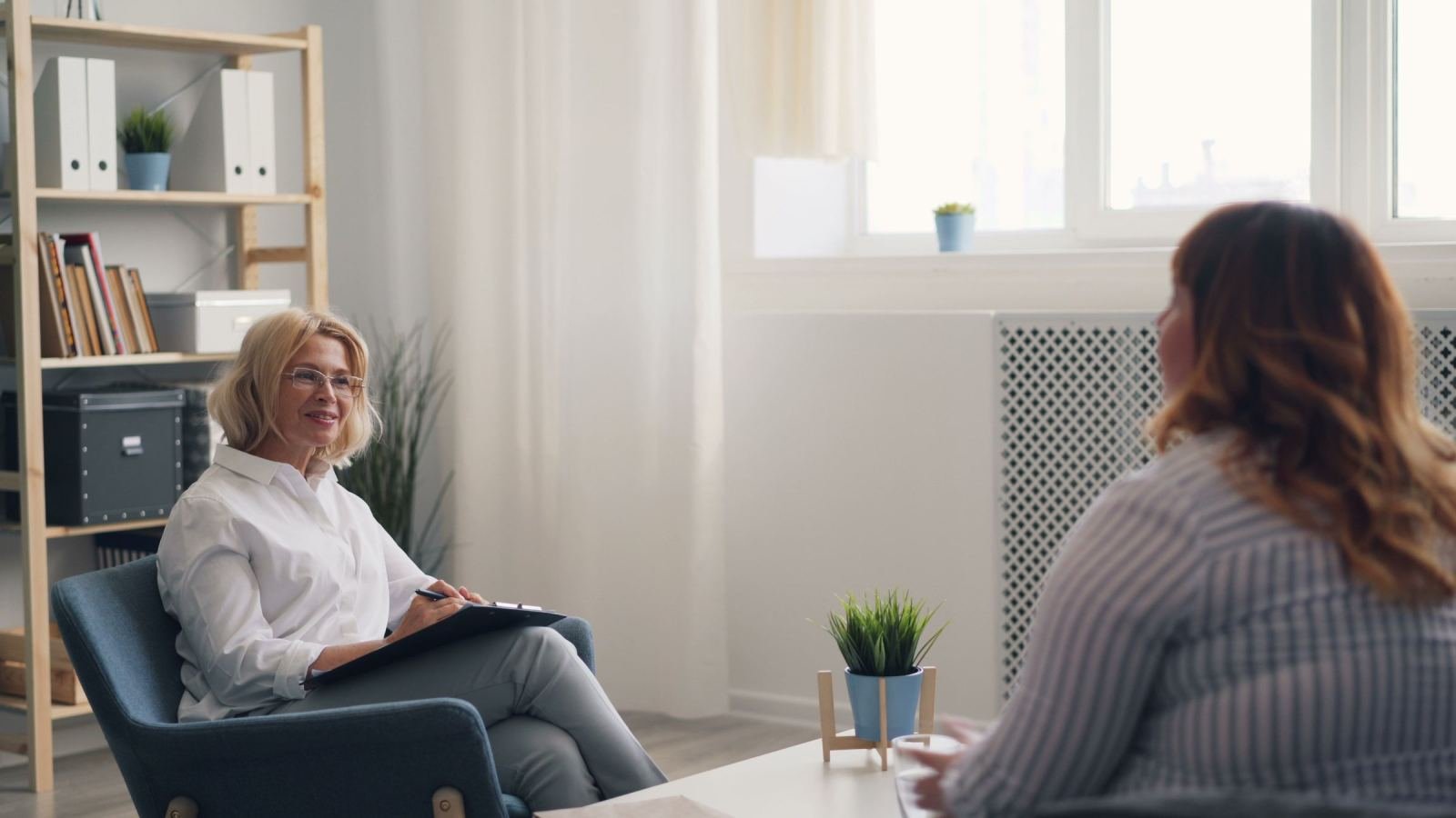 A therapist holding a clipboard speaks with a client in a well-lit room.