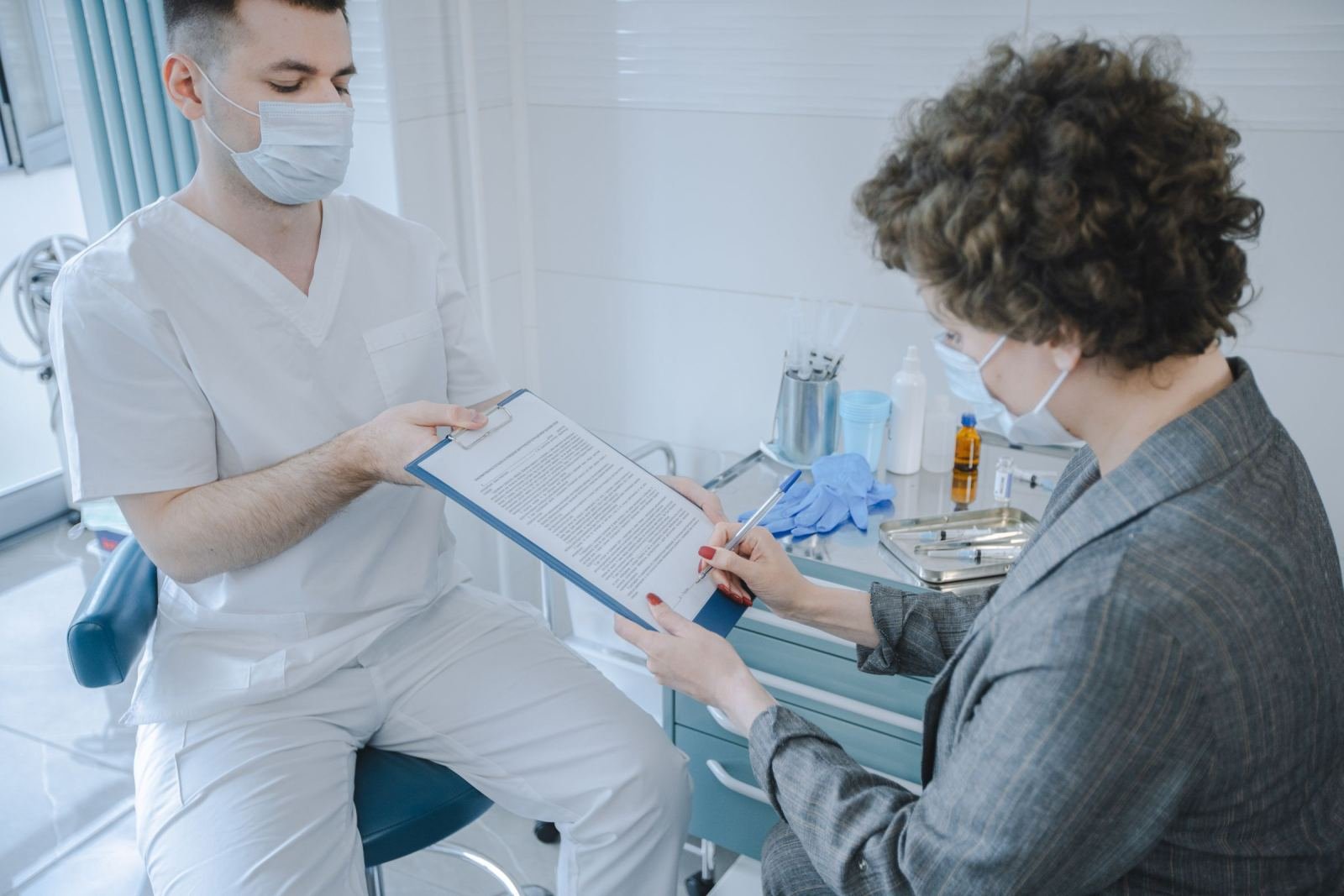 A clinician in scrubs and a mask reviews a document with a woman in a grey blazer