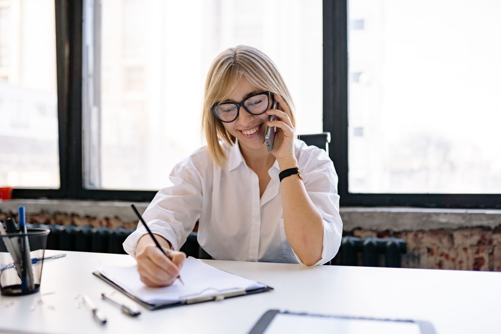 A woman wearing glasses sits at a desk, talking on the phone and writing in a notebook.