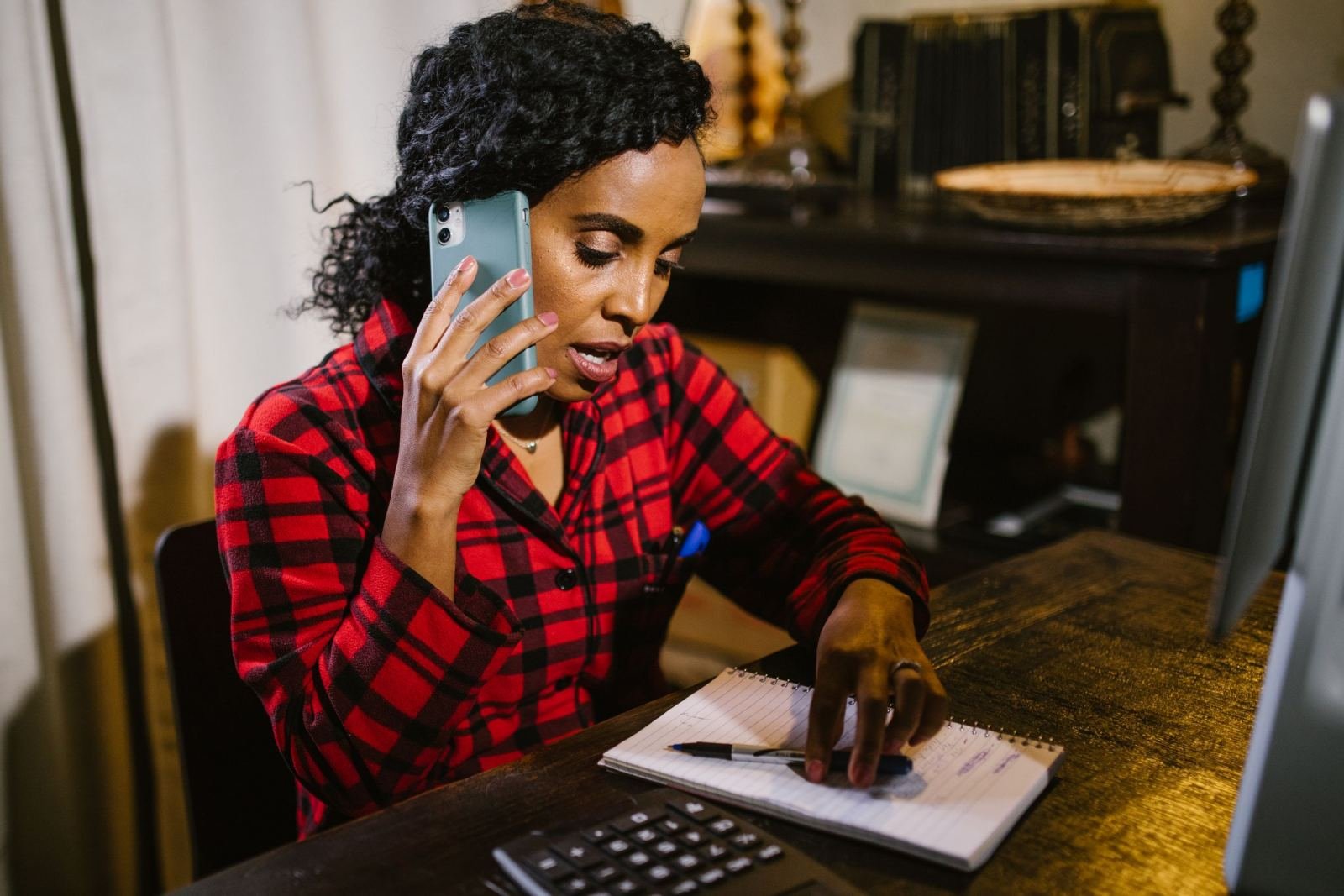 A woman uses a smartphone while she writes notes at her desk.