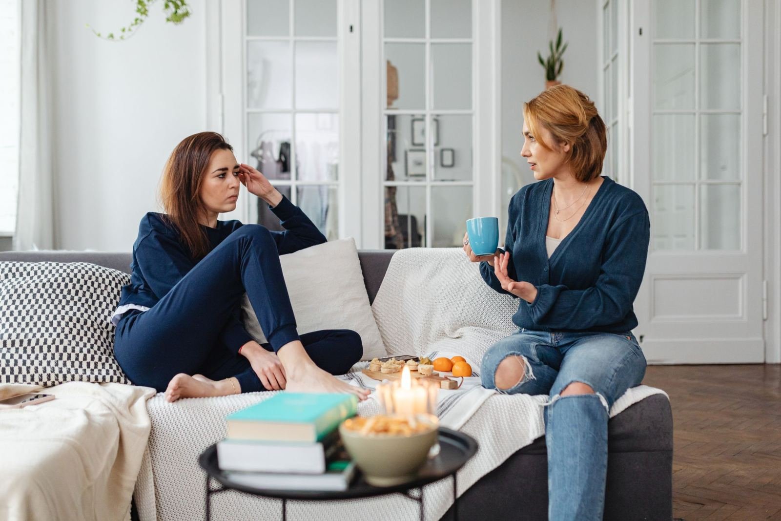 Two women talk while sitting on a couch. One of them holds a blue mug.
