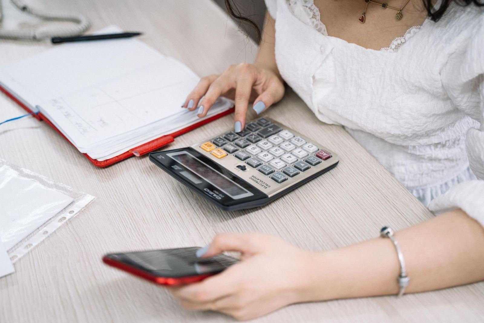 A person using a calculator and a smartphone at a desk with a notebook