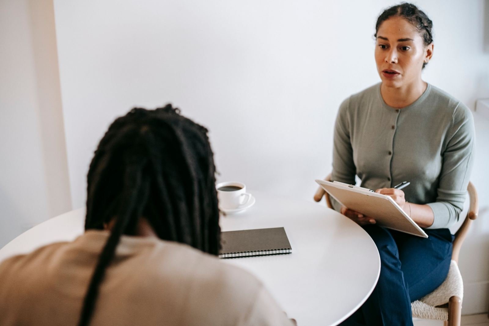 A clinician with a clipboard talks to a client at a white round table